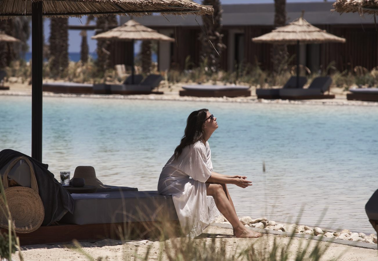 Woman sitting in the shade at the edge of a beautiful lagoon at Casa Cook North Coast wearing a white flowing cover up and sunglasses