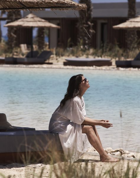Woman sitting in the shade at the edge of a beautiful lagoon at Casa Cook North Coast wearing a white flowing cover up and sunglasses