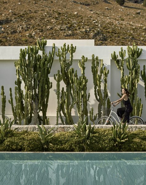 Woman cycling past cactus-lined suites at Casa Cook Rhodes, surrounded by minimalist architecture and mountain views in a peaceful adults-only retreat in Kolymbia