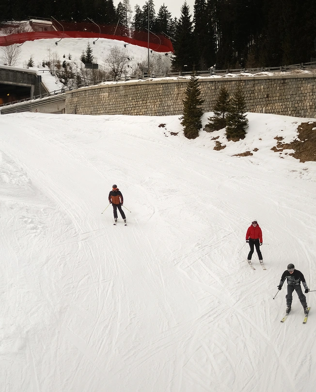 Three skiers descend a freshly groomed piste in Madonna di Campiglio, viewed from above, with pine trees and a stone retaining wall lining the slope.