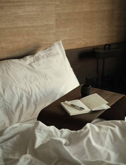 An open journal and pen rest on a dark walnut bedside shelf beside a matte black ceramic cup, against rumpled white linen bedding and a travertine-textured headboard wall at Casa Cook Madonna di Campiglio.