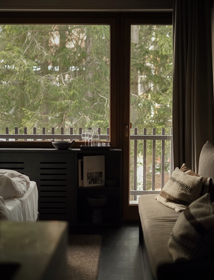 A moody guest room corner at Casa Cook Madonna, with floor-to-ceiling windows framing snow-dusted pine trees, a linen sofa scattered with woven cushions, and a water carafe on a dark side table.