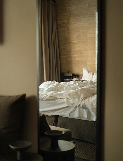 A floor-length mirror reflects a softly lit Casa Cook hotel bedroom — rumpled white bedding, linen curtains, a travertine feature wall, and small dark stools in the foreground.