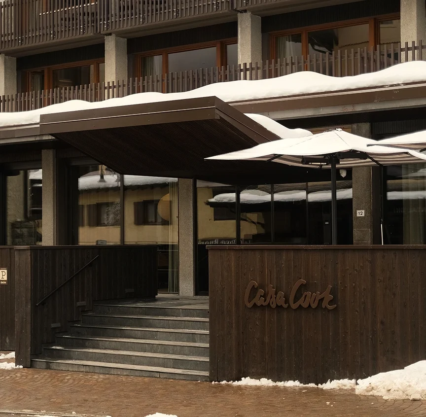 The exterior entrance of Casa Cook Madonna di Campiglio in winter — dark timber cladding, the Casa Cook logo in bronze lettering, stone steps, and snow-dusted parasols framed by the hotel's balconied facade.