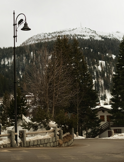 A quiet Alpine street corner in Madonna di Campiglio in winter, with an ornate lamp post, pine trees, and a snow-covered mountain peak rising in the background.