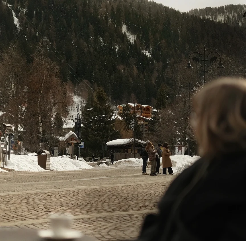 A woman with blonde hair sits with a coffee cup in the foreground, gazing out across a snow-dusted cobblestone piazza in Madonna di Campiglio, with alpine chalets and pine-forested slopes beyond.