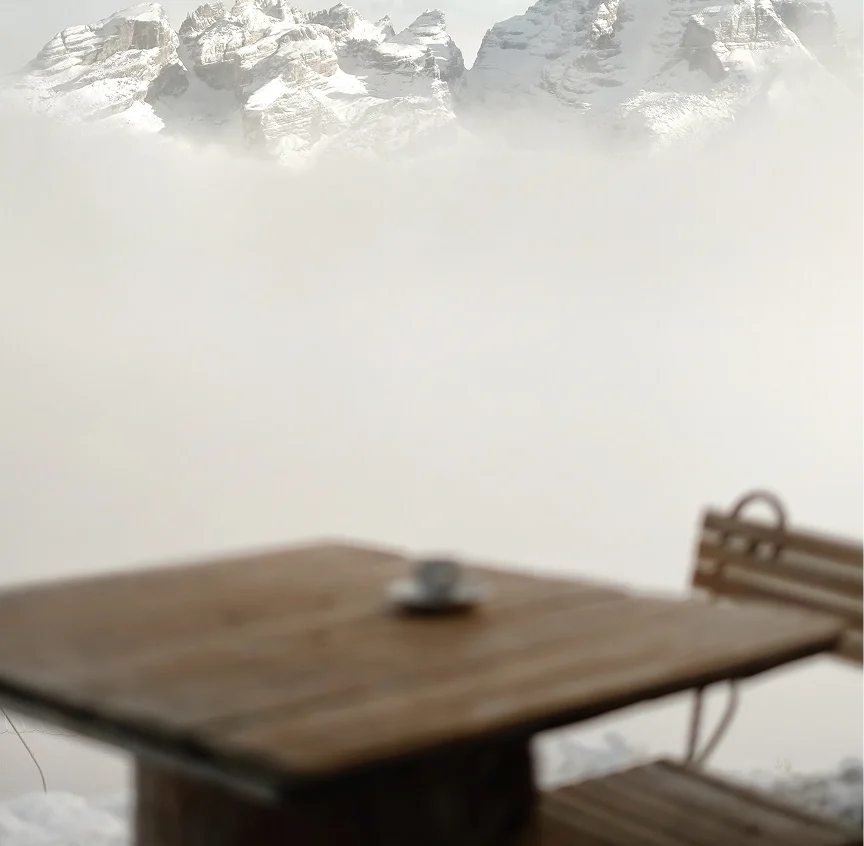 A wooden outdoor table with a lone espresso cup sits in the foreground, out of focus, while snow-capped Dolomite peaks pierce dramatically through a swirling sea of cloud above.
