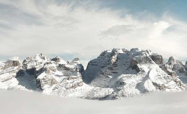 A panoramic view of the Dolomites in winter — jagged, snow-capped peaks emerging dramatically above a thick sea of cloud under a pale blue sky.