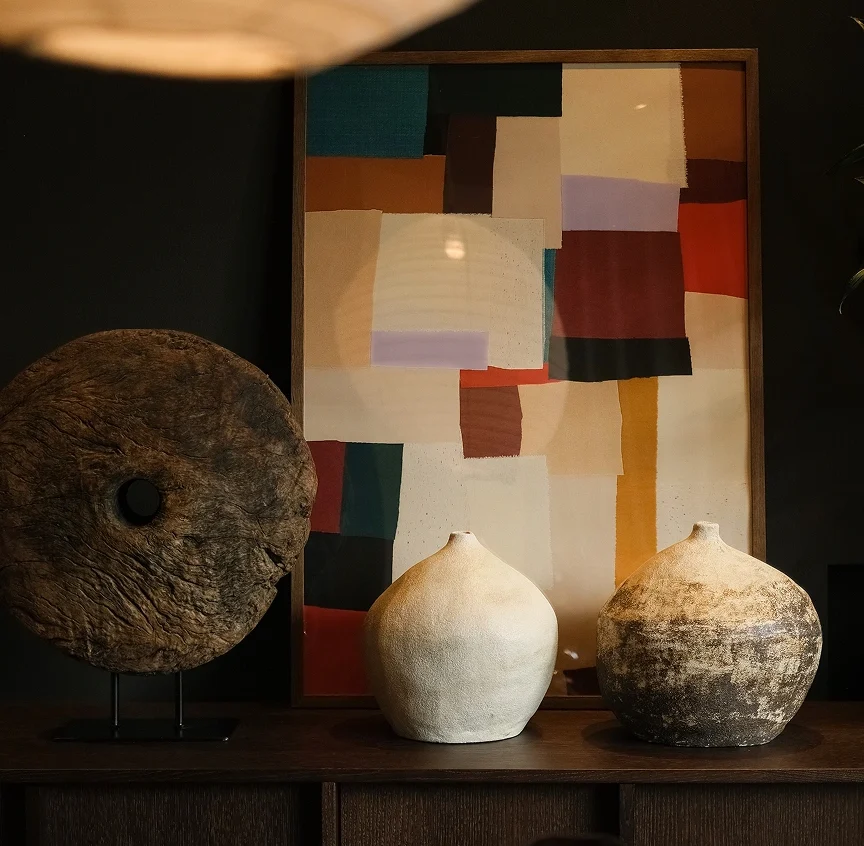 A vignette of carefully curated objects on a dark wood console at Casa Cook Madonna: two organic stoneware vases, a weathered circular wooden sculpture on a stand, and a bold abstract artwork in earthy tones of teal, red, ochre and lavender.