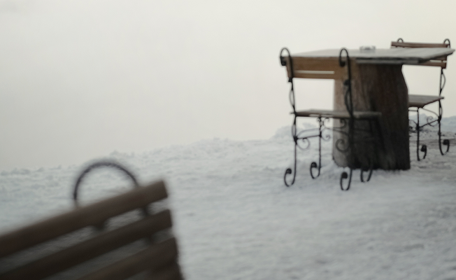 A snow-blanketed outdoor terrace in the Italian Alps, with a wrought iron bistro table and wooden bench half-buried in fresh snow, evoking stillness after a heavy snowfall.