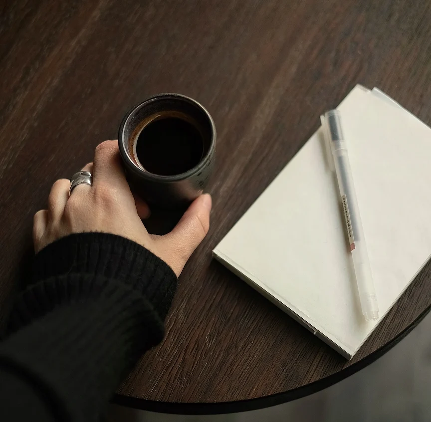 Overhead close-up of a ringed hand cradling a matte black ceramic cup of black coffee on a dark walnut table, with an open notebook and pen beside it — slow morning energy at Casa Cook.