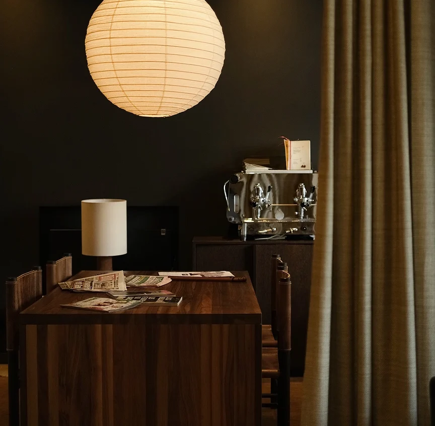 Inside Casa Cook Madonna's café space — a glowing paper lantern pendant hangs above a dark walnut communal table scattered with newspapers, beside a professional espresso machine and floor-length linen curtain.