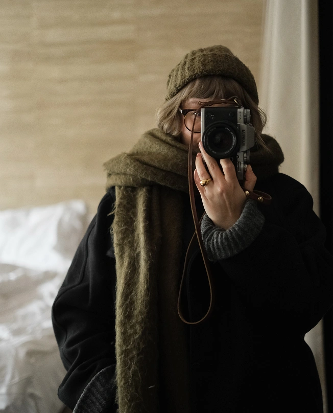 A self-portrait reflected in a hotel mirror: the photographer, wearing a chunky olive knit beanie, oversized scarf, and black coat, holds a vintage film camera up to her face against a warm travertine wall.