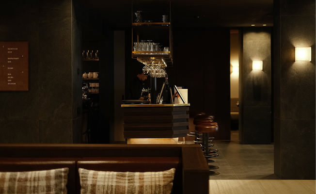 The dimly lit bar and reception area of Casa Cook Madonna di Campiglio, with dark stone walls, warm pendant lighting, a professional espresso machine on a brass-accented counter, and leather bar stools.