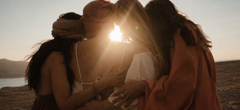 Four women standing together with their arms around each other looking out to the sunset with their backs to the camera